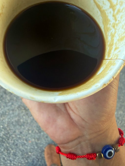 Close-up photo of a hand holding a cup filled with a dark liquid (coffee or similar drink); the wrist wears a red string bracelet with a blue evil eye charm.