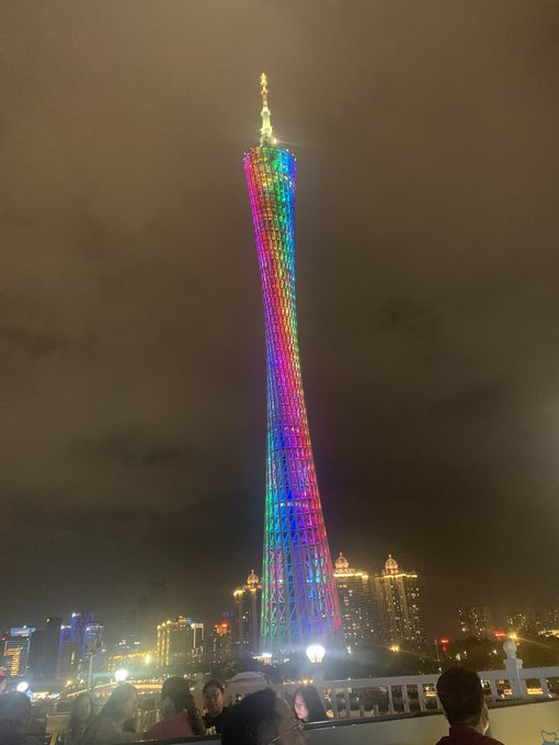 A night photograph of the Canton Tower in Guangzhou illuminated with rainbow-colored LED lights, with crowds of people and the city skyline visible at the base.