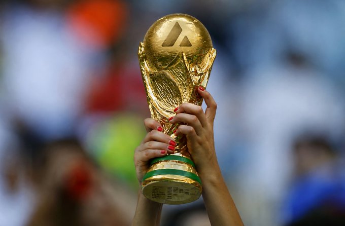 Hands with red nail polish hold up the golden FIFA World Cup trophy against a blurred stadium background.