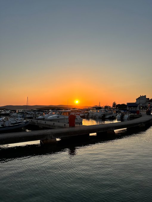 A serene sunset over a harbor with boats docked along piers, where calm waters reflect the golden hues of the setting sun. The image's tranquil scene contrasts with the post's anticipation for a directional move, suggesting a "calm before the storm" as the market awaits a narrative shift.