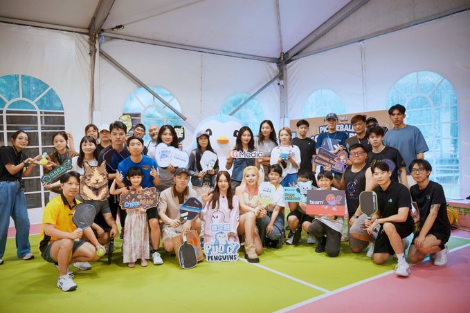 A large group photo of people posing together at a pickleball event under a white tent, holding signs with logos including 'Medio', 'Pickleball', and 'Penguins'.