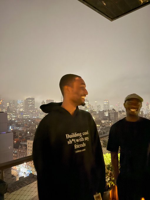 Two smiling young men pose on a rooftop at night with a city skyline behind them; the man on the left wears a black hoodie reading "Building cool sh*t with my friends."