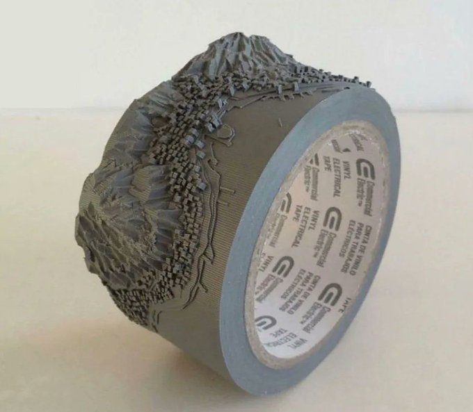 A sculptural artwork made from a roll of gray tape, with the tape peeled and cut to form a detailed miniature mountain landscape on top of the roll.
