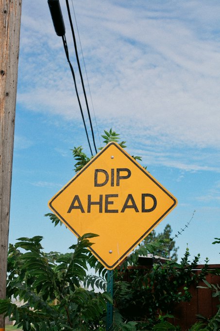 Yellow diamond-shaped road sign reading 'DIP AHEAD' mounted on a post, surrounded by green foliage under a partly cloudy sky.