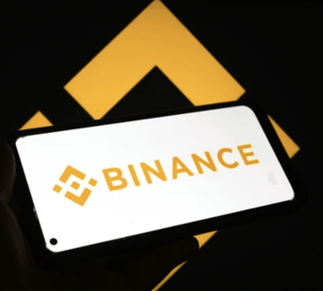 The image features a person holding up their phone displaying the Binance logo. The phone screen shows the word "BINANCE" written in yellow letters against a black background. The text appears to be related to an event or news, possibly about Bitcoin or its wallet.