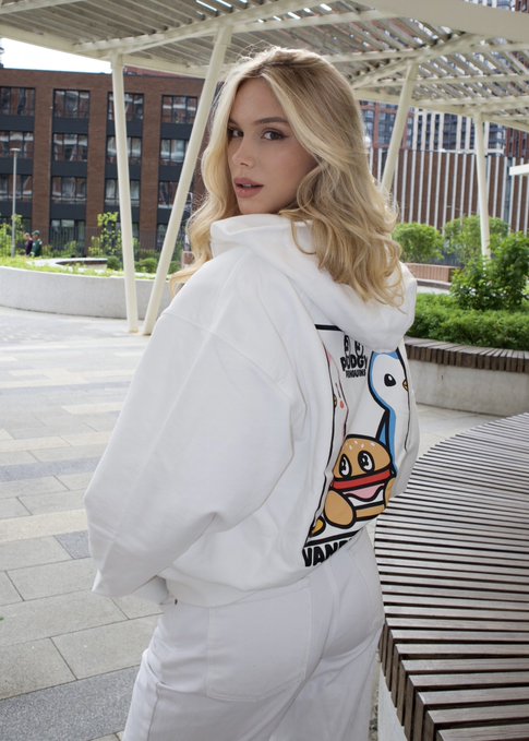 The image features a woman wearing a white hoodie that has a cartoon character design of a dog on it. She appears to be posing for a photo while standing next to a wooden bench, which suggests she might be at an outdoor event or gathering. The background includes buildings and trees, indicating the setting could be in an urban area with some greenery nearby. The image also contains text that reads "GM Wall Street may close, but markets keep moving." This message seems to be related to the content of the post about investing in US stocks and commodities.
