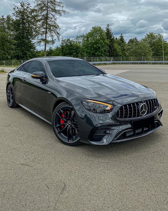 A dark gray Mercedes-AMG coupe parked on asphalt with a cloudy sky and trees in the background.