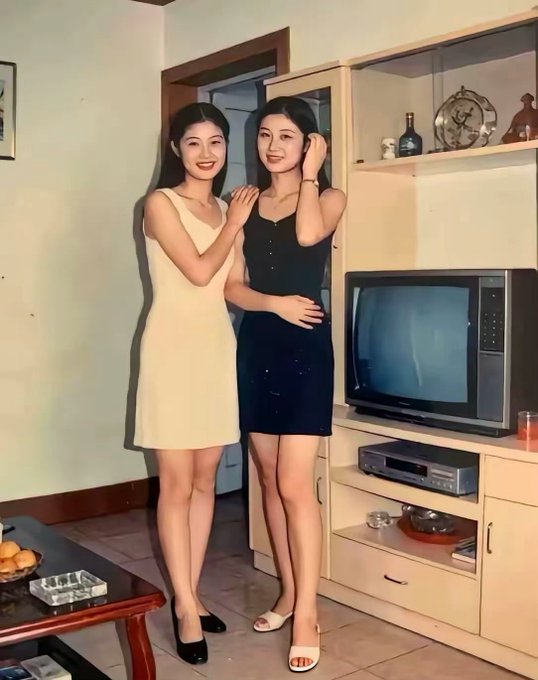 A vintage-style photograph of two smiling young women in dresses posing together in a living room with a retro television set.