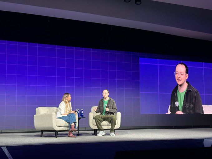 The image shows a man and woman sitting on chairs facing each other with their backs to the camera, likely participating in an event or panel discussion. They are positioned behind a screen that displays a video of another person speaking. The background features a blue wall adorned with white squares, creating a visually appealing backdrop for the speakers. The caption below the image reads "INSIGHT: “The Twitter vibes are really negative, but when it comes to adoption of the technology, it's kinda good".