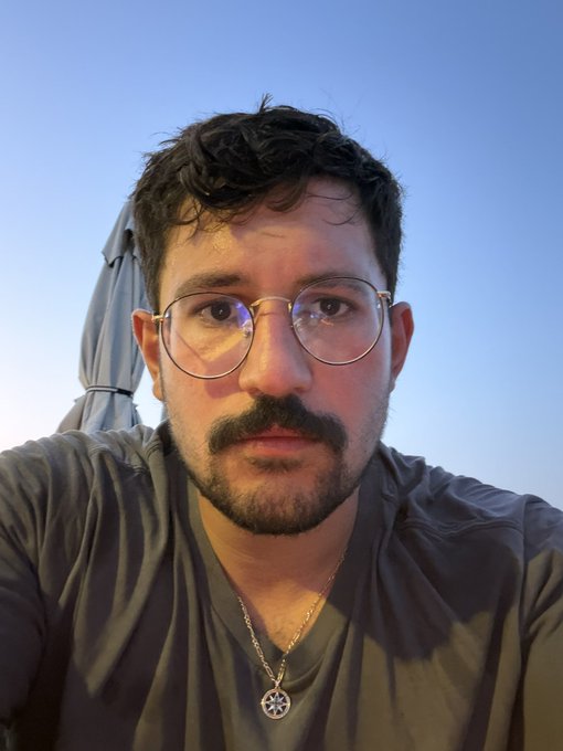 A man with curly dark hair, glasses, and a mustache poses for a selfie after an intense boxing workout, wearing a gray t-shirt and a peace sign necklace. The caption invites followers to tag a Monad KOL to fight, humorously suggesting he's tired and looking for a playful showdown after his training session.