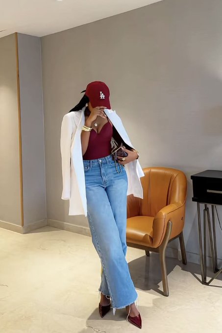 A woman wearing an LA Dodgers cap, burgundy top, white blazer, and blue jeans posing indoors near an orange chair.