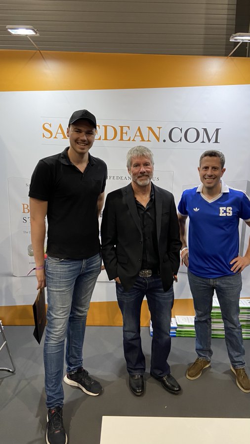 The image features three men standing next to each other at a booth for the Saadean.com cryptocurrency exchange, with one of them holding a book titled "The Bitcoin Book". The booth has a sign that says "Saadean.com" on it, and there are stacks of books visible in the background. One man is wearing a black shirt while the others have blue shirts. They appear to be posing for a photo together at the booth.