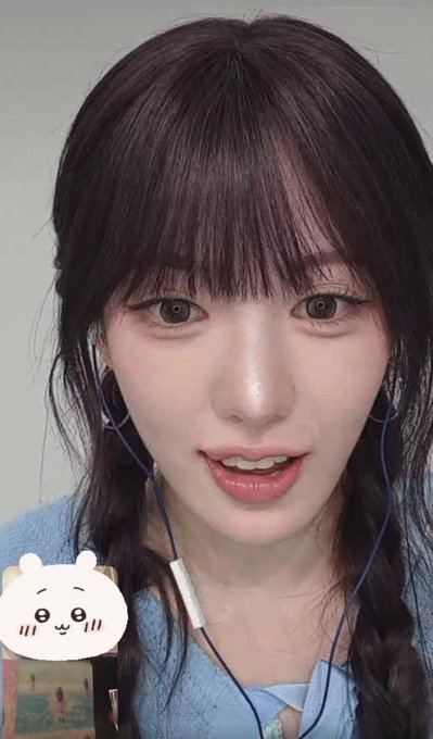 A close-up selfie shows a young woman with dark hair in two braids, bangs, wired earphones, and a light blue top; a small white cartoon sticker sits near the lower left.