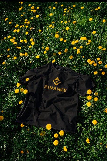 A black t-shirt with the yellow Binance logo and text lies on grass surrounded by yellow dandelion flowers.