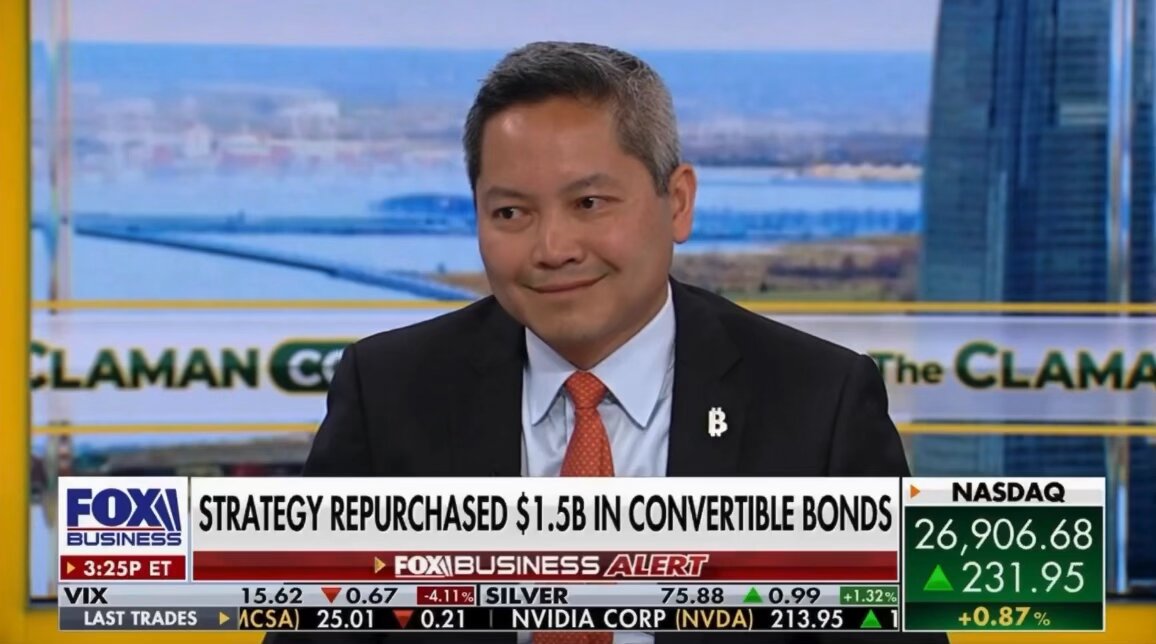 The image shows a man wearing a suit and tie sitting at a desk, with a TV screen behind him displaying news about Bitcoin and financial markets. The headline reads "Strategy CEO Phong Le made statements about the company's agenda during a program he participated in". This suggests that the man is discussing or analyzing the current state of Bitcoin and its impact on the financial market.