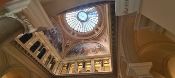 An upward view of an ornate building interior featuring a domed stained-glass ceiling, painted frescoes, and columns.