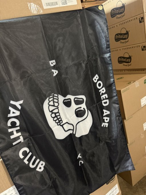 A black Bored Ape Yacht Club flag featuring the iconic white skull logo, laid out among cardboard boxes labeled "FritoLay Good fun!"