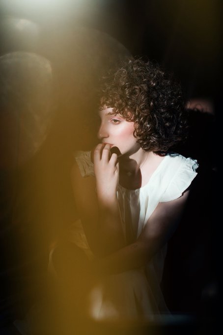 A young woman with curly hair and dramatic lighting appears in a contemplative pose, hand near her face. The moody, artistic portrait complements the accompanying post announcing a new 1/1 NFT art piece collaboration with @zahraladani on Objkt for 500 xtz, celebrating #TezosTuesday.