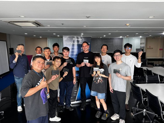 A group of people pose together in a classroom or meeting room, each holding up what appears to be a Tangem hardware wallet, with a Tangem-branded banner visible in the background displaying Thai text ("กระเป๋าเงินฮาร์ดแวร์" - meaning "hardware wallet"). The image captures the successful conclusion of Tangem's first-ever Thai-language lecture on self-custody held at Chulalongkorn University, with participants proudly displaying their new wallets.