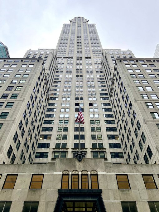 A dramatic upward-angled photograph of a tall, ornate Art Deco skyscraper with a grid-like facade of windows, an American flag flying near the entrance, and intricate stone detailing at the lower levels.
