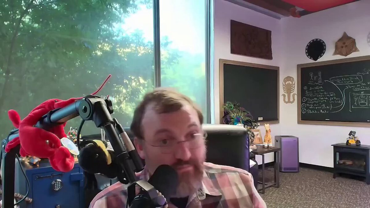 The image shows a man wearing glasses sitting behind a microphone at a desk, likely participating in an online radio show or podcast. The room features a window with blinds that allow natural light into the space, creating a comfortable atmosphere for the host's recording session. The background includes various objects such as potted plants and a chair, adding to the overall ambiance of the room.