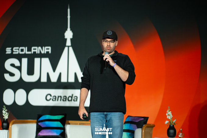 A man wearing glasses, a black cap, and a black sweater speaks into a microphone on stage at the 'SOLANA SUMMIT Canada' event.
