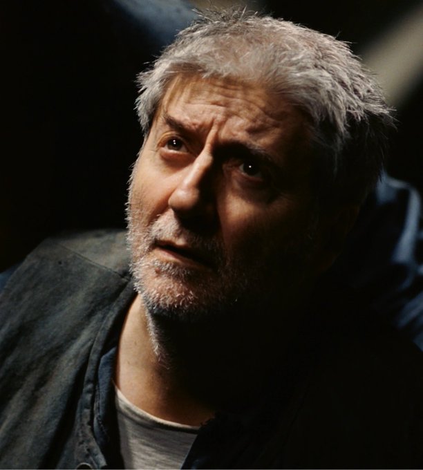A close-up portrait of a middle-aged man with gray hair and a beard, looking off to the side with a serious expression against a dark background.