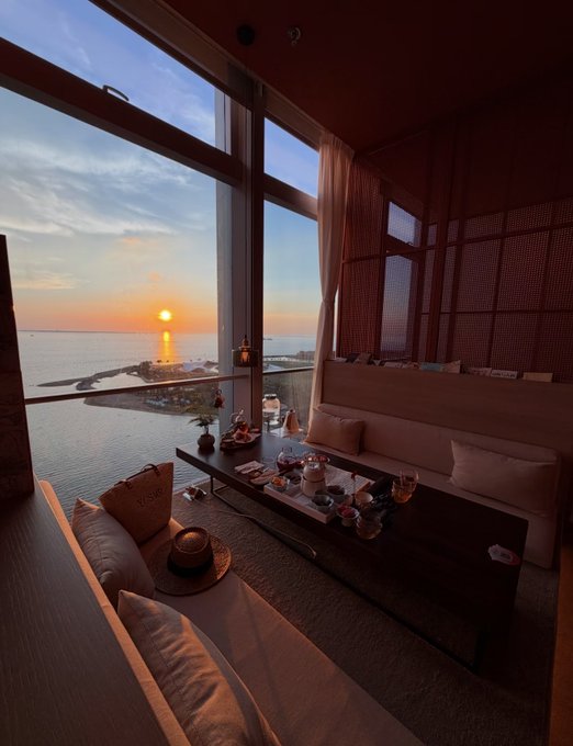An interior view of a room with a sunset over the ocean seen through large floor-to-ceiling windows, featuring a seating area with a tea set arranged on a low table.