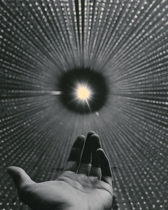 A black and white photograph of a hand reaching toward a radiant light source surrounded by radiating dotted patterns, creating a cosmic effect.