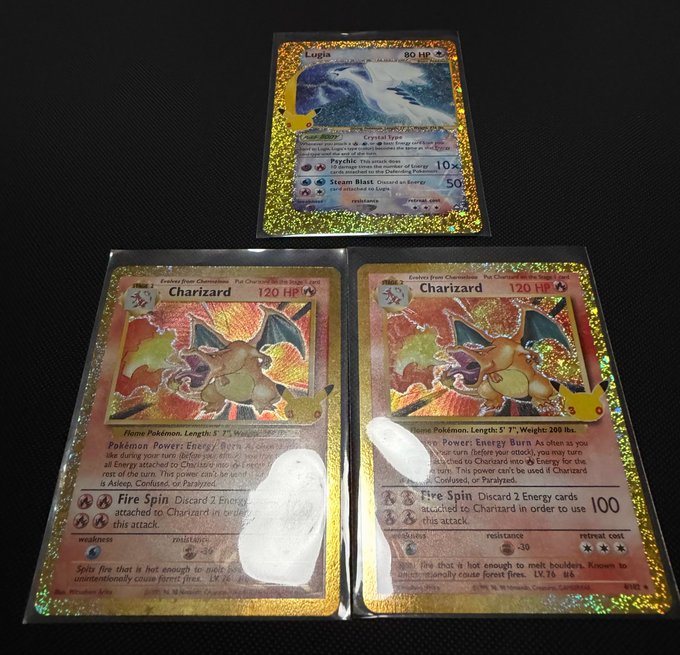 Three holographic Pokémon cards displayed on a dark surface, including one Lugia card (80 HP) and two Charizard cards (120 HP), both featuring the "Fire Spin" attack.