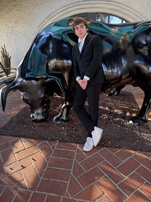 A young man in a black suit and white sneakers leans against a large bronze bull statue on a brick patio.