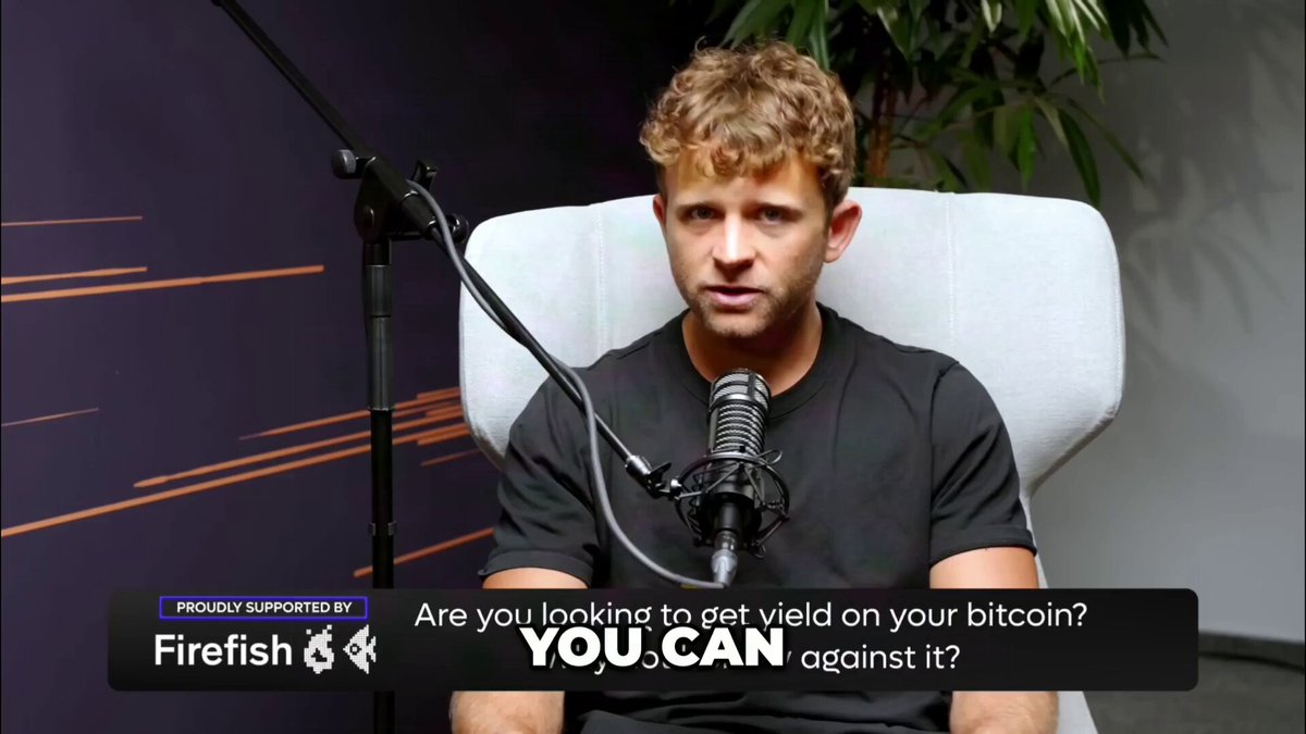The image features a man sitting in a chair with a microphone attached to his shirt, suggesting that he might be hosting or participating in an interview or podcast discussion about Bitcoin. The background of the image shows a purple wall, which could be part of the studio setup for the show where this conversation is taking place.