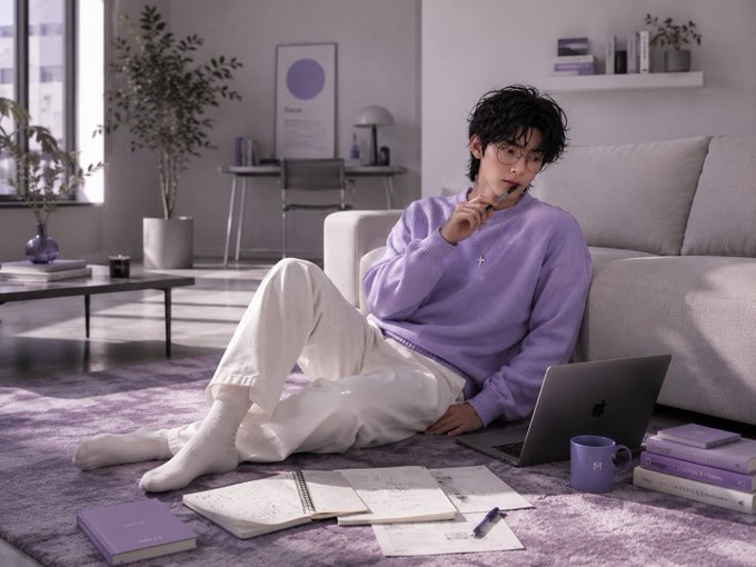 A man wearing glasses and a lavender sweater sits on the floor of a modern living room, working on an Apple laptop with notebooks scattered around him.