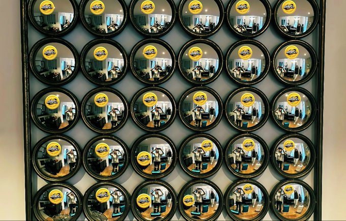 The image features a wall with numerous circular mirrors arranged in rows, creating an interesting visual effect. The mirrors are reflecting various images of people, including some wearing backpacks or carrying handbags. These reflections give the impression that there are multiple copies of the same person and their belongings, possibly symbolizing the concept of "many faces" or "many stories." This unique arrangement of mirrors can be seen as a creative way to explore different perspectives and experiences in a single image.