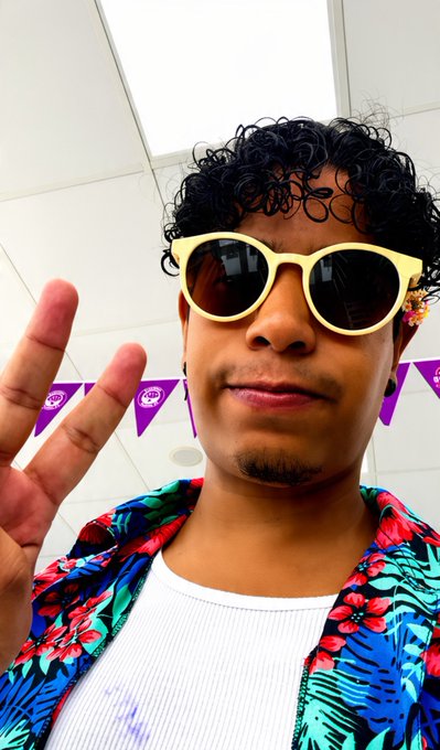 A selfie of a person with curly black hair wearing yellow sunglasses, a white tank top, and a colorful floral shirt, making a peace sign.
