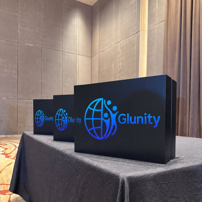 Three black boxes with blue illuminated "Glunity" globe logos sit on a draped table in a conference room setting.