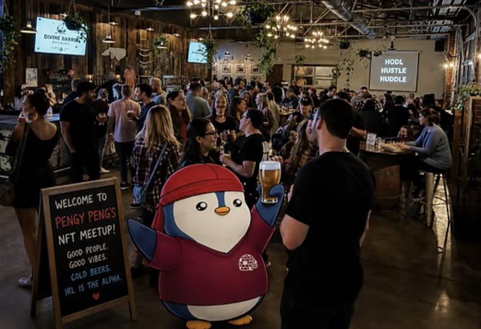 A crowded indoor event venue with a 'Pengy Peng's NFT Meetup!' welcome sign and a cartoon penguin mascot holding a beer mug.
