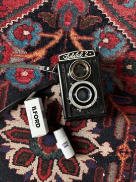 A vintage black "Mirex 22" twin-lens reflex camera sits on an ornate red, blue, and black patterned rug alongside an "ILFORD" film box and other small accessories.