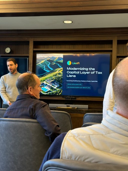 The image shows two men sitting in chairs in front of a large screen television displaying an advertisement for a company called "LFI" or "Capital Layer of Tax". The man on the left side of the room appears to be giving a presentation, while the other man is attentively watching and listening. Both individuals are wearing ties, which suggests they might have professional roles in the context of the presentation.