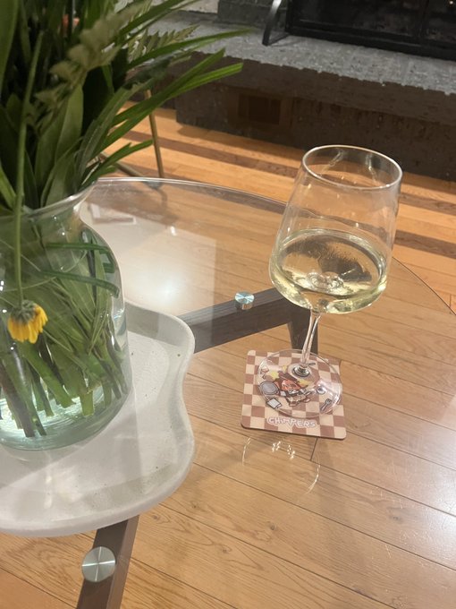 A glass of white wine sits on a 'Croptober' coaster next to a vase of daisies and ferns on a wooden table with a glass top.