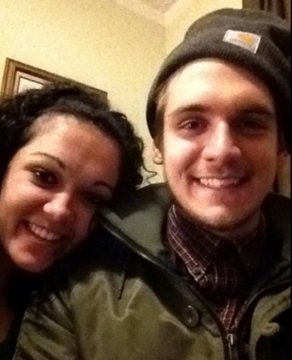 A close-up selfie of a smiling couple indoors, with a woman having dark curly hair and a man wearing a dark Carhartt beanie and a green jacket.