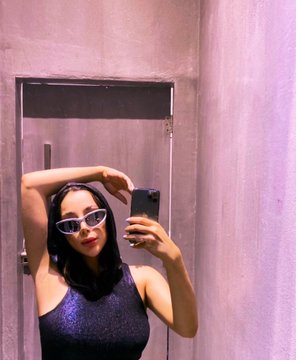 A woman wearing white sunglasses and a sparkly black top takes a mirror selfie in what appears to be a fitting room, posing with one hand raised near her head in a stylish summer-ready pose that fits the "Summer" theme of the QT Art and Artist series.