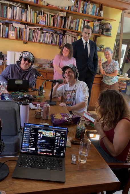 A group of people gathered around a table in a room with bookshelves, recording a podcast with microphones and a laptop visible.