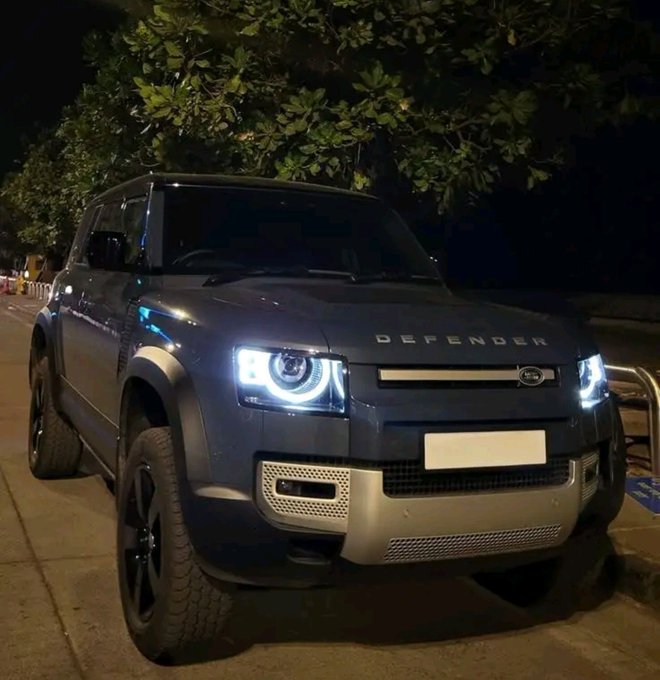 A dark blue Land Rover Defender is parked at night with its headlights on; “DEFENDER” is visible across the hood.