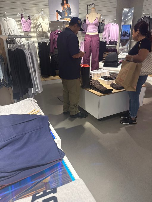 Inside a clothing store, a man examines items on a display table while a woman holding a Lululemon shopping bag stands nearby.