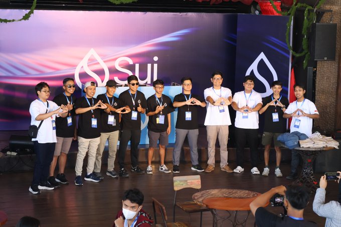 A group photo of young men posing on stage in front of a 'Sui' branded backdrop, making a hand gesture, with a Vietnamese flag visible on the right side.