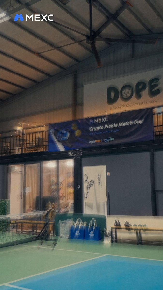 The image shows a tennis court with a blue net dividing the two halves. There are several bags placed around the court, including handbags near the net and another one further away from the net. The scene also includes a banner hanging on the wall of the tennis court, which reads "DOPE" in white letters.