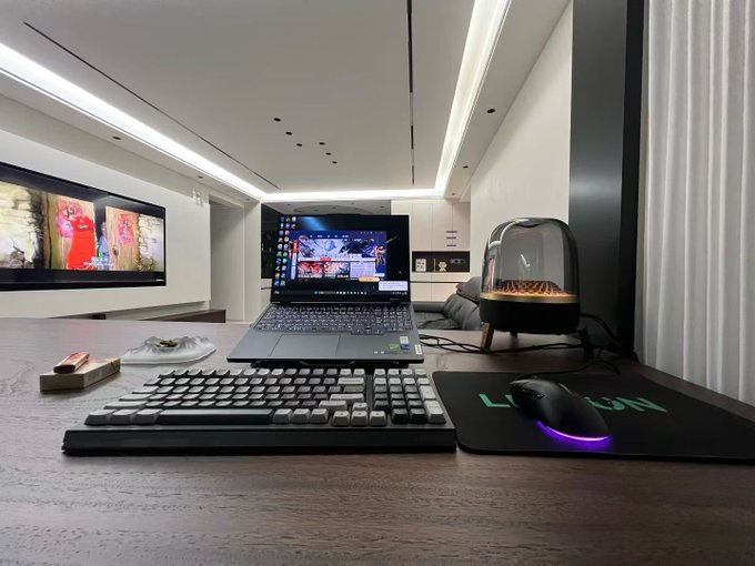 A minimalist desk setup with a laptop displaying colorful wallpapers, a mechanical keyboard, a gaming mouse on a 'LIN' mousepad, and a modern interior with recessed ceiling lighting.