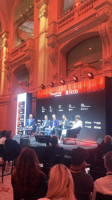 The image shows a group of people sitting around a stage, with a large screen behind them displaying information about cryptocurrency. The audience appears engaged and interested in the topic being discussed. The text "Proofoftalk" suggests that this event might be related to blockchain technology or digital currencies. The room is filled with chairs where attendees are seated, and there's a person wearing a tie visible on one of the individuals.