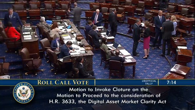 A high-angle view of the U.S. Senate floor shows senators engaged in conversation and seated at their desks during a roll call vote on H.R. 3633, the Digital Asset Market Clarity Act, to invoke cloture on the motion to proceed, with an on-screen graphic displaying the time as 7:14 and the vote as an unofficial tally.